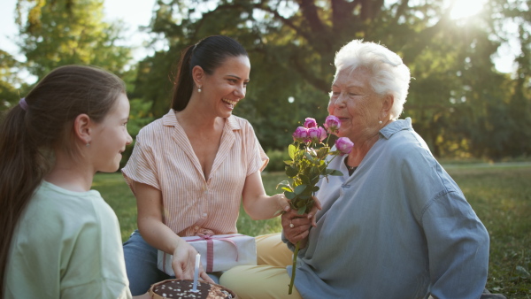 A small girl with mother giving cake and flowers to grandmother in a park, celebrating birthday.