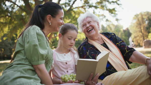 A small girl with mother and grandmother resting in a park, reading a book.