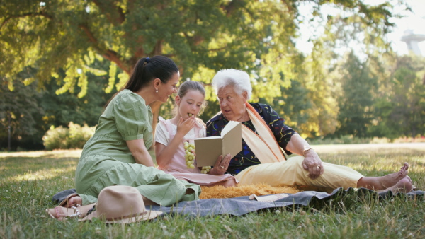 A small girl with mother and grandmother resting in a park, reading a book.