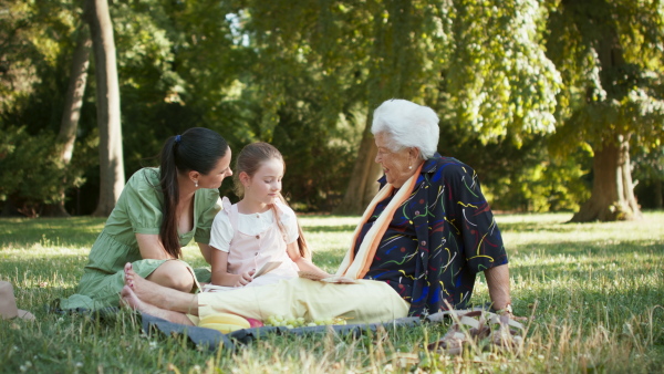 A small girl with mother and grandmother resting in a park, reading a book.