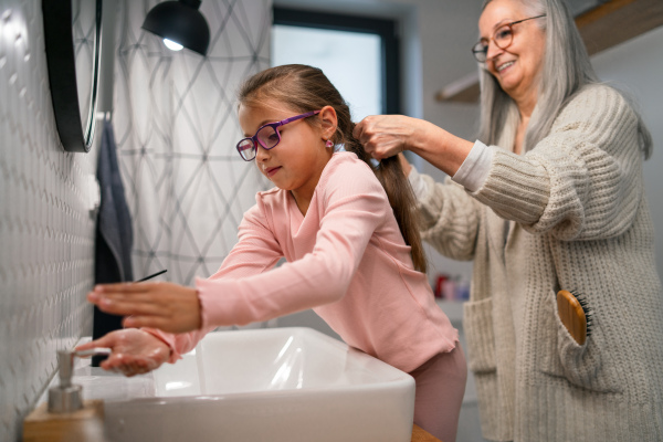 A senior grandmother and granddaughter standing indoors in bathroom, daily routine concept.