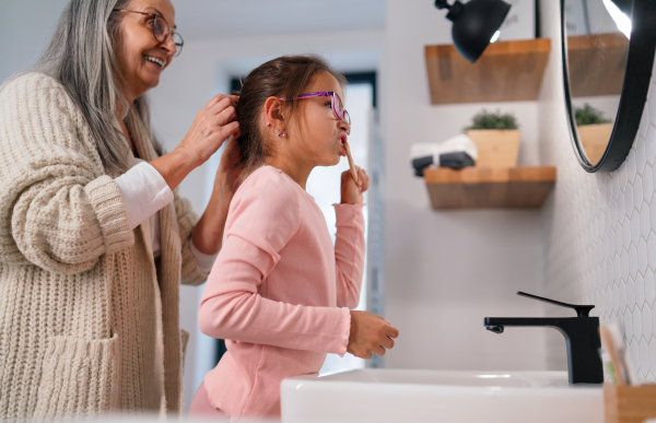 A senior grandmother making a ponytail to her granddaughter indoors in bathroom in the morning.