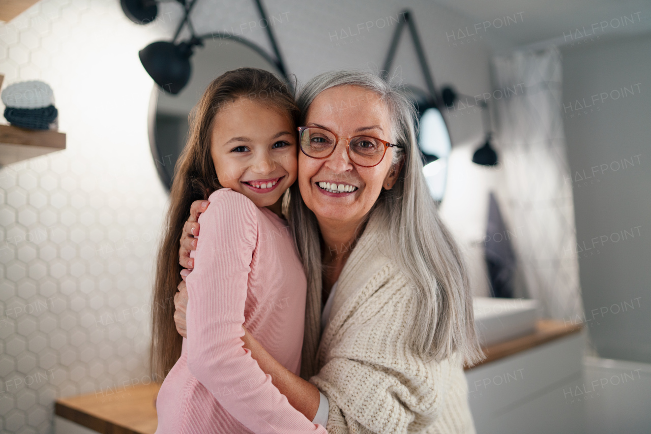 A senior grandmother and granddaughter standing indoors in bathroom, hugging and looking at camera.