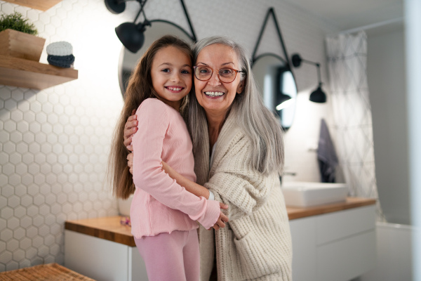 A senior grandmother and granddaughter standing indoors in bathroom, hugging and looking at camera.