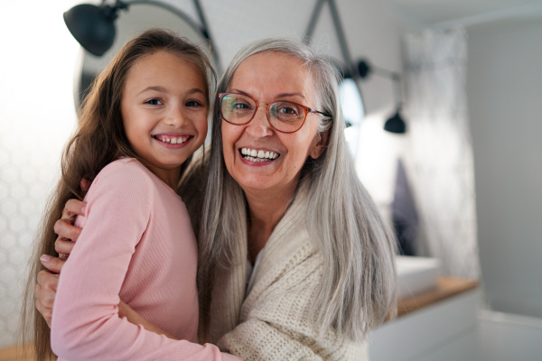 A senior grandmother and granddaughter standing indoors in bathroom, hugging and looking at camera.