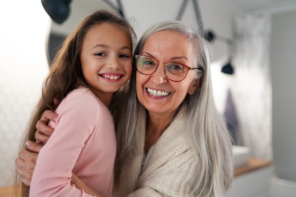 A senior grandmother and granddaughter standing indoors in bathroom, hugging and looking at camera.