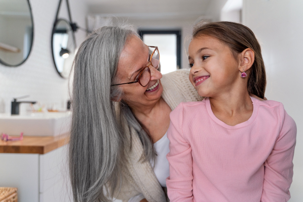 A senior grandmother and granddaughter standing indoors in bathroom, looking at each other.