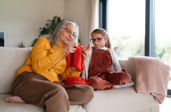 A little girl sitting on sofa with her grandmother and learning to knit indoors at home.