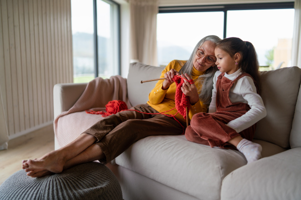 A little girl sitting on sofa with her grandmother and learning to knit indoors at home.