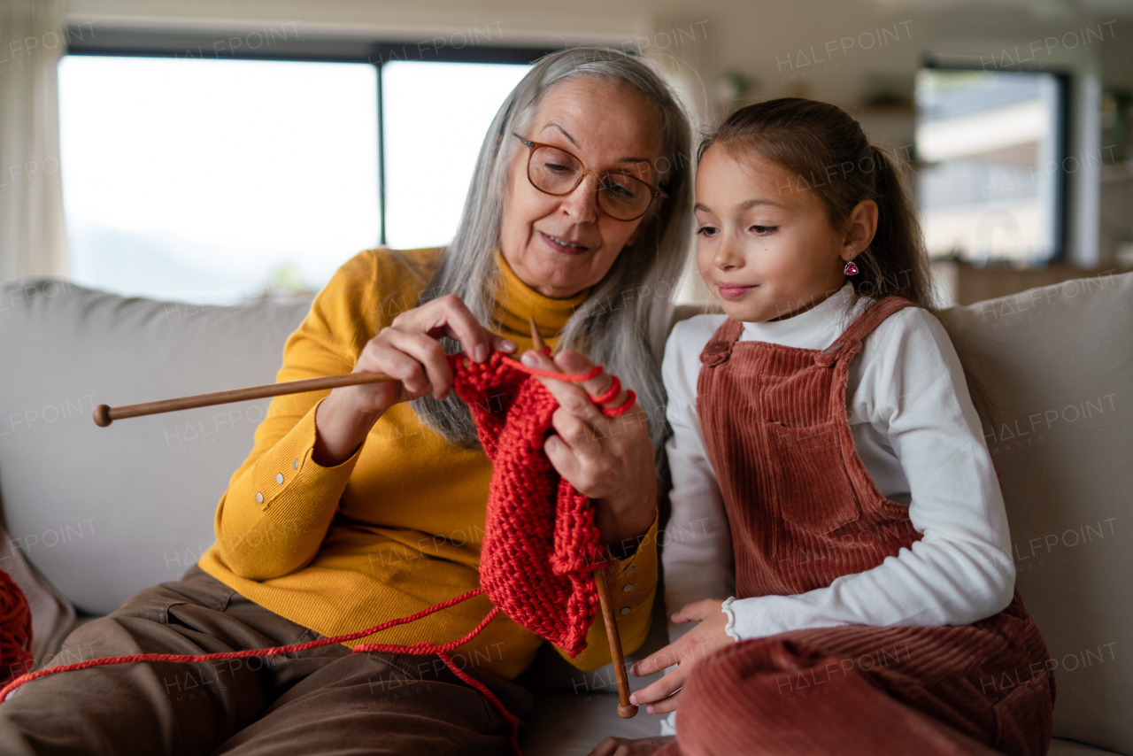 A little girl sitting on sofa with her grandmother and learning to knit indoors at home.