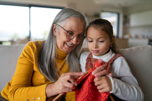 A little girl sitting on sofa with her grandmother and learning to knit indoors at home.