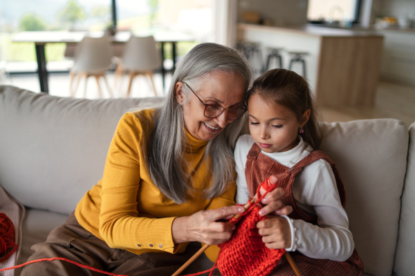 A little girl sitting on sofa with her grandmother and learning to knit indoors at home.