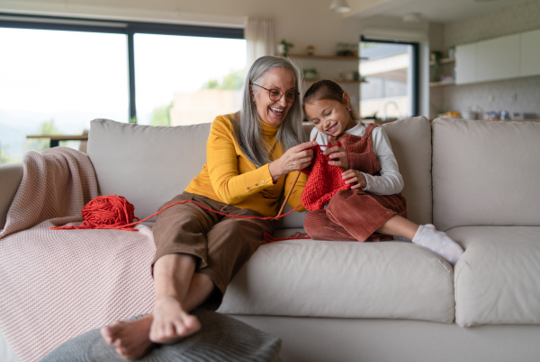 A little girl sitting on sofa with her grandmother and learning to knit indoors at home.