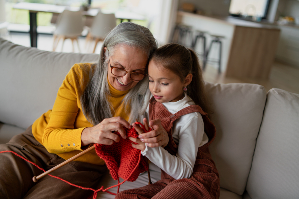 A little girl sitting on sofa with her grandmother and learning to knit indoors at home.