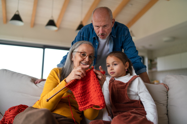 A little girl sitting on sofa with her grandparents and learning to knit indoors at home.