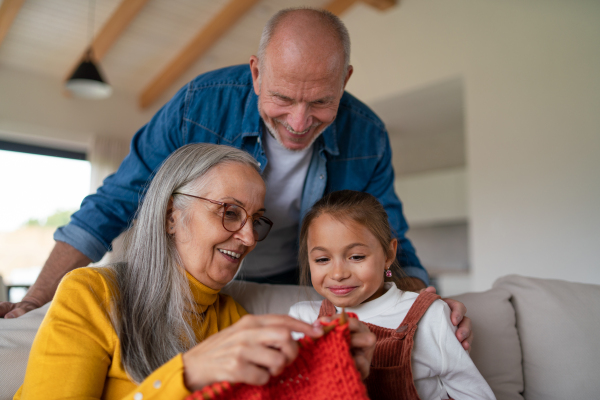 A little girl sitting on sofa with her grandparents and learning to knit indoors at home.