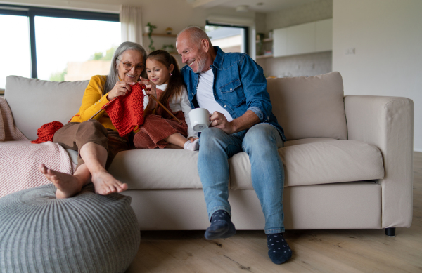 A little girl sitting on sofa with her grandparents and learning to knit indoors at home.