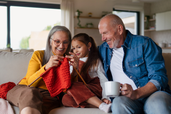 A little girl sitting on sofa with her grandparents and learning to knit indoors at home.