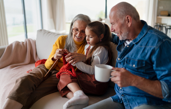 A little girl sitting on sofa with her grandparents and learning to knit indoors at home.