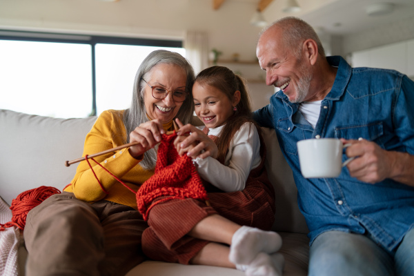 A little girl sitting on sofa with her grandparents and learning to knit indoors at home.