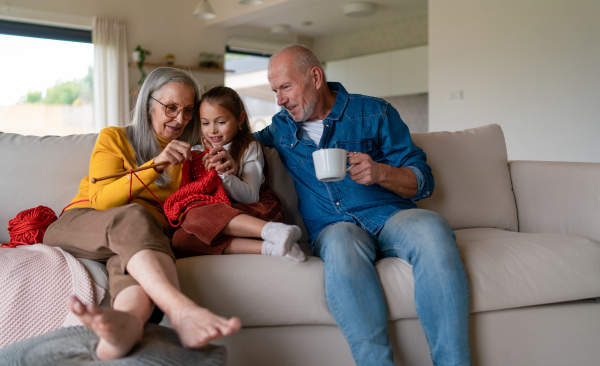 A little girl sitting on sofa with her grandparents and learning to knit indoors at home.