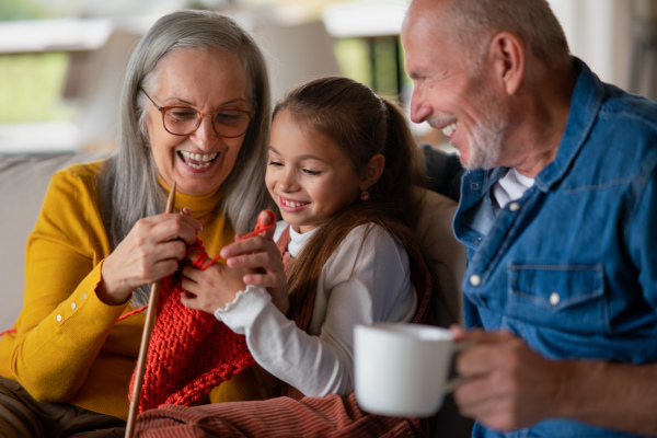 A little girl sitting on sofa with her grandparents and learning to knit indoors at home.