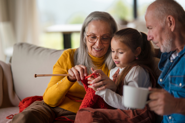 A little girl sitting on sofa with her grandparents and learning to knit indoors at home.