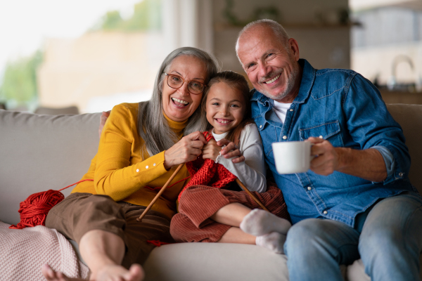 A little girl sitting on sofa with her grandparents and learning to knit indoors at home.