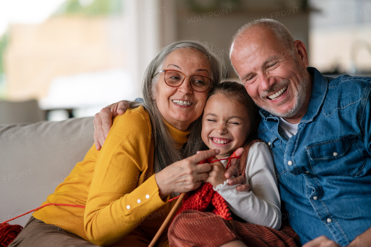 A little girl sitting on sofa with her grandparents and learning to knit indoors at home.