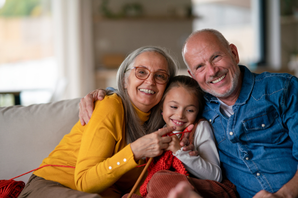 A little girl sitting on sofa with her grandparents and learning to knit indoors at home.