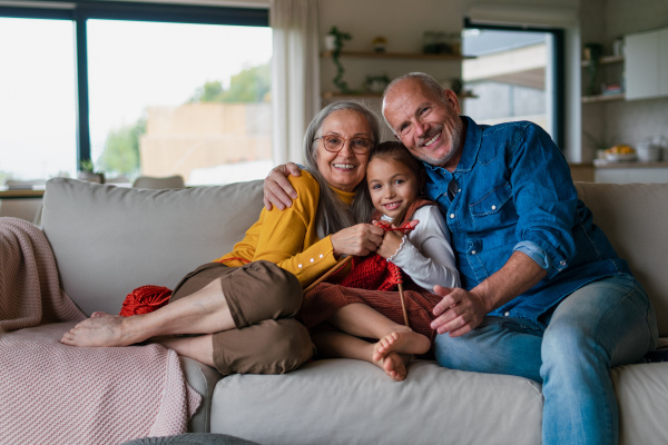 A little girl sitting on sofa with her grandparents and learning to knit indoors at home, looking at camera.