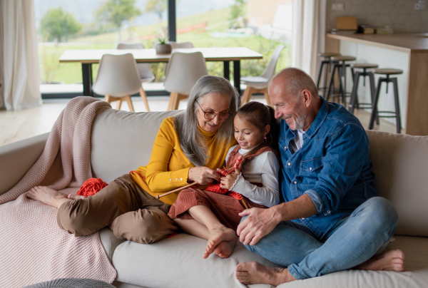 A little girl sitting on sofa with her grandparents and learning to knit indoors at home.