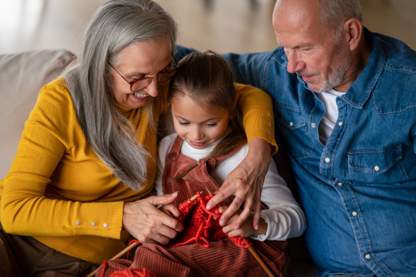 A little girl sitting on sofa with her grandparents and learning to knit indoors at home.