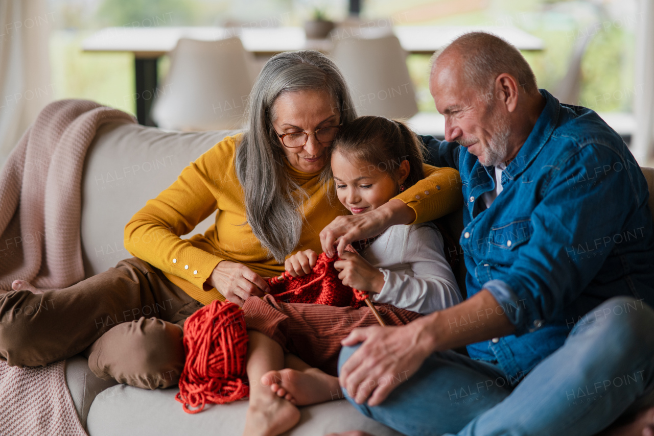 A little girl sitting on sofa with her grandparents and learning to knit indoors at home.