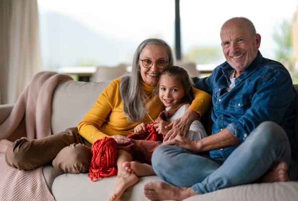 A little girl sitting on sofa with her grandparents and learning to knit indoors at home.