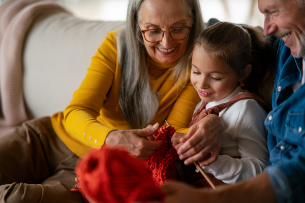 A little girl sitting on sofa with her grandparents and learning to knit indoors at home.