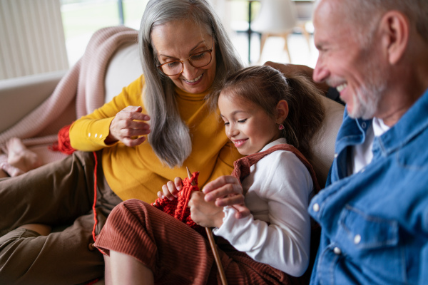 A little girl sitting on sofa with her grandparents and learning to knit indoors at home.