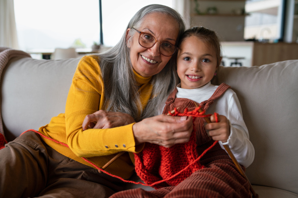 A little girl sitting on sofa with her grandmother and learning to knit indoors at home.