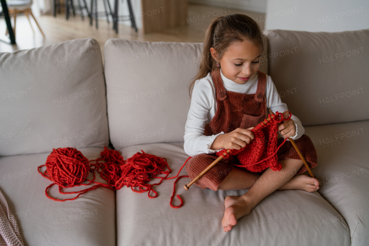 A little girl sitting on sofa and learning to knit indoors at home.