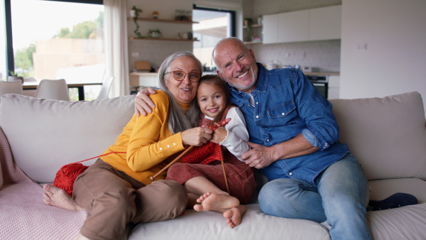 A little girl sitting on sofa with her grandparents and learning to knit indoors at home.