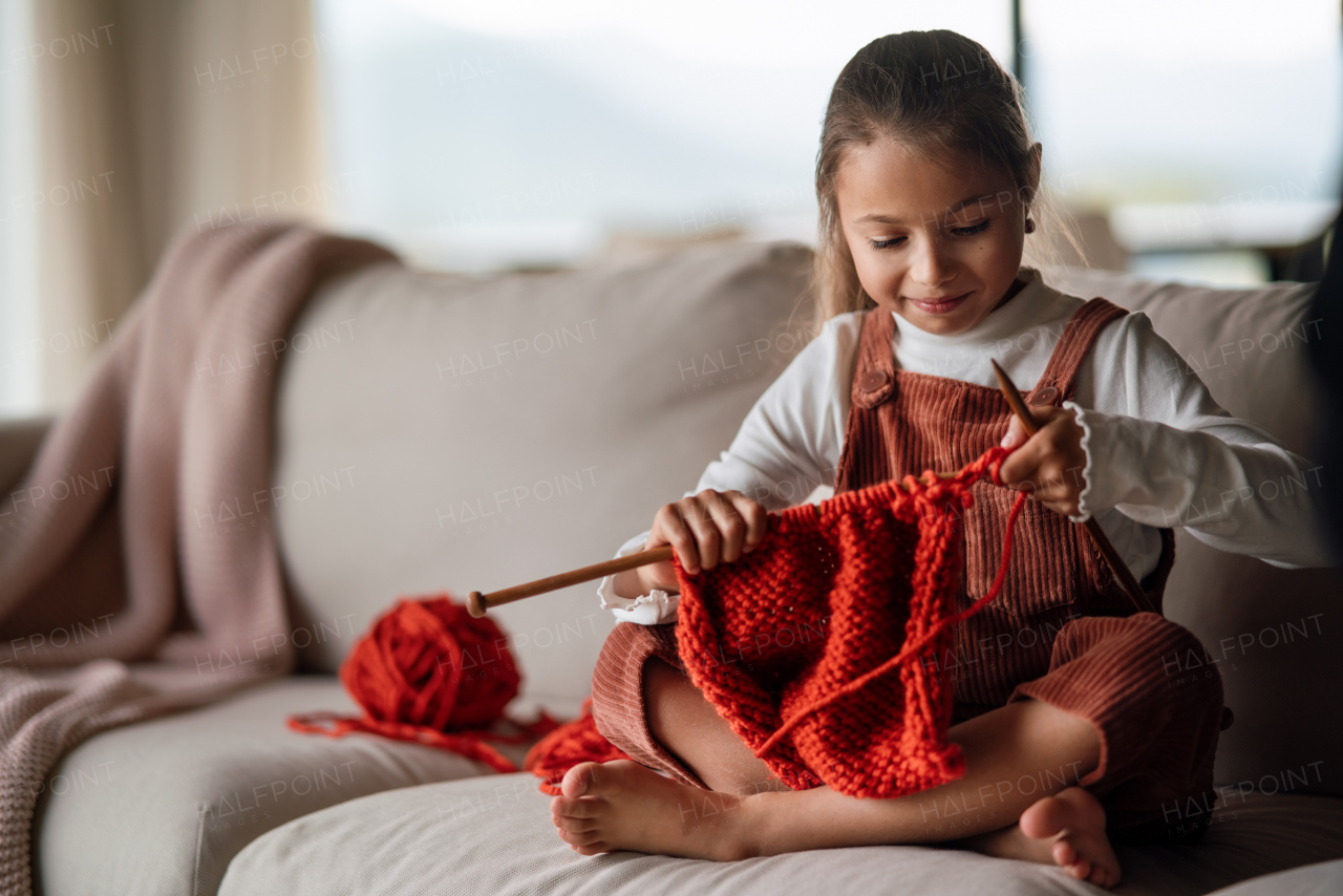 A little girl sitting on sofa and learning to knit indoors at home.