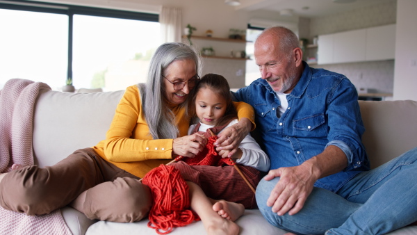 A little girl sitting on sofa with her grandparents and learning to knit indoors at home.