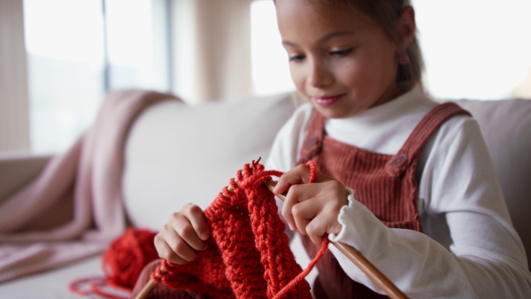 A little girl sitting on sofa and learning to knit indoors at home.