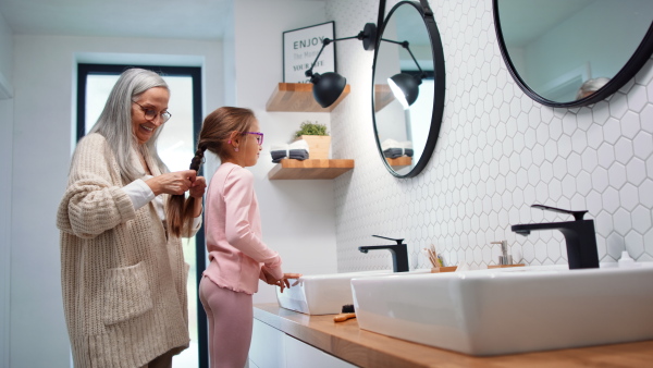 A senior grandmother making a plait to her granddaughter indoors in bathroom in the morning.