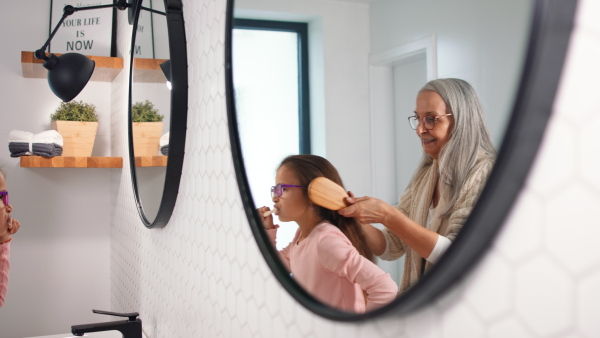 A senior grandmother and granddaughter standing indoors in bathroom, combing hair in morning.