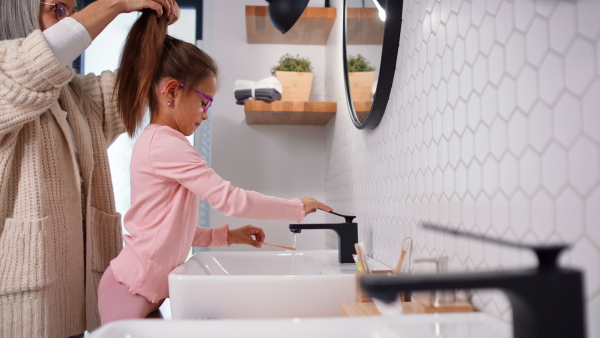 A senior grandmother making a ponytail to her granddaughter indoors in bathroom in the morning.