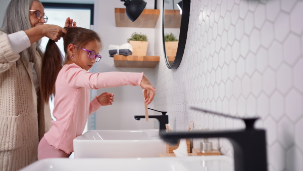 A senior grandmother and granddaughter standing indoors in bathroom, brusing teeth and hair in morning