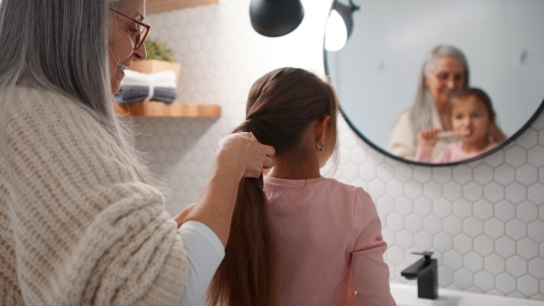 A senior grandmother and granddaughter standing indoors in bathroom, brusing teeth and hair in morning