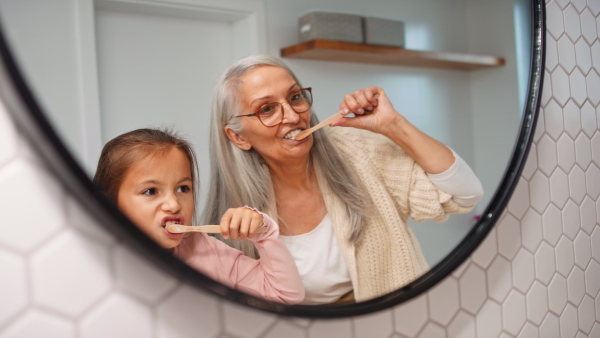 A senior grandmother with granddaughter standing indoors in bathroom, brusing teeth and looking at mirror.