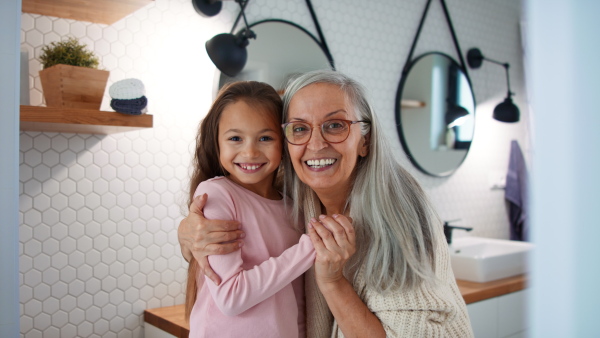 A senior grandmother and granddaughter standing indoors in bathroom, hugging and looking at camera.